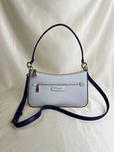 Load image into Gallery viewer, A light blue pebbled leather handbag with navy blue leather straps and silver-tone hardware, displayed on a draped white fabric background. The brand name 'kate spade' is visible on the front metal plaque.